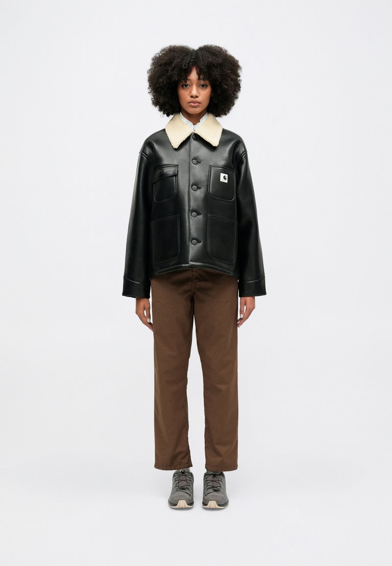 Czarna kurtka z sztucznej skóry z beżowym futrzanym kołnierzem, dużymi kieszeniami z przodu i zapięciem na guziki, w połączeniu z brązowymi spodniami i szarymi butami.