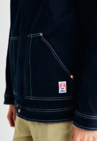 Giacca in denim blu scuro con cuciture bianche, grandi tasche frontali e un'etichetta Wrangler sulla tasca sinistra. Accenti in hardware di rame visibili.