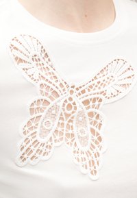 Top blanc avec un grand motif papillon en dentelle centré sur la poitrine, montrant des motifs complexes et des espaces ouverts.