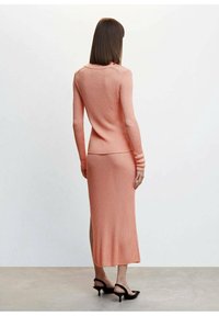 Robe longue à côtes en corail doux, ajustée au corsage, avec des manches longues et une fente sur le côté. Associée à des sandales à talons noires.
