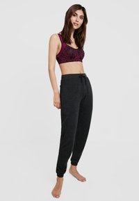 Femme debout pieds nus, portant un soutien-gorge de sport bordeaux à motifs et un pantalon de jogging gris foncé avec cordon, devant un fond uni.