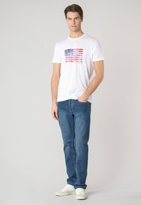 T-shirt bianco a maniche corte con una grafica sbiadita della bandiera americana sul davanti, abbinato a jeans blu e sneakers bianche.