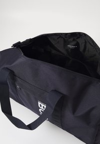 Fekete sporttáska textil külsővel, felső cipzárral és párhuzamos fogantyúkkal. A belső részen sima felület és márkajelzés található.