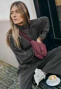 Giacca oversized grigia a righe sottili sovrapposta a un dolcevita scuro, abbinata a una borsa a tracolla bordeaux e sneakers bianche. Una tazza di caffè è posata nelle vicinanze.