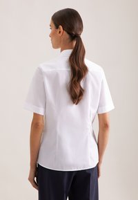 Weißes kurzärmeliges Hemd mit einem strukturierten Kragen, glatter Textur und seitlichen Schlitzen, kombiniert mit dunkelblauen Hosen. Einfaches und sauberes Design.