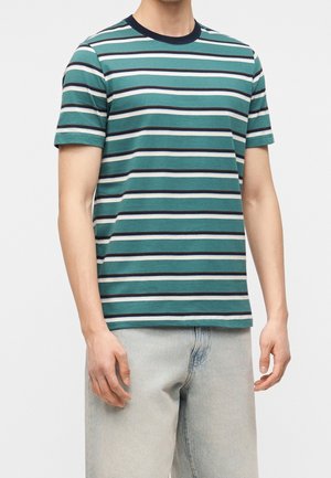 Homme portant un t-shirt à manches courtes rayé bleu sarcelle, noir et blanc avec un jean gris clair, debout devant un fond uni.