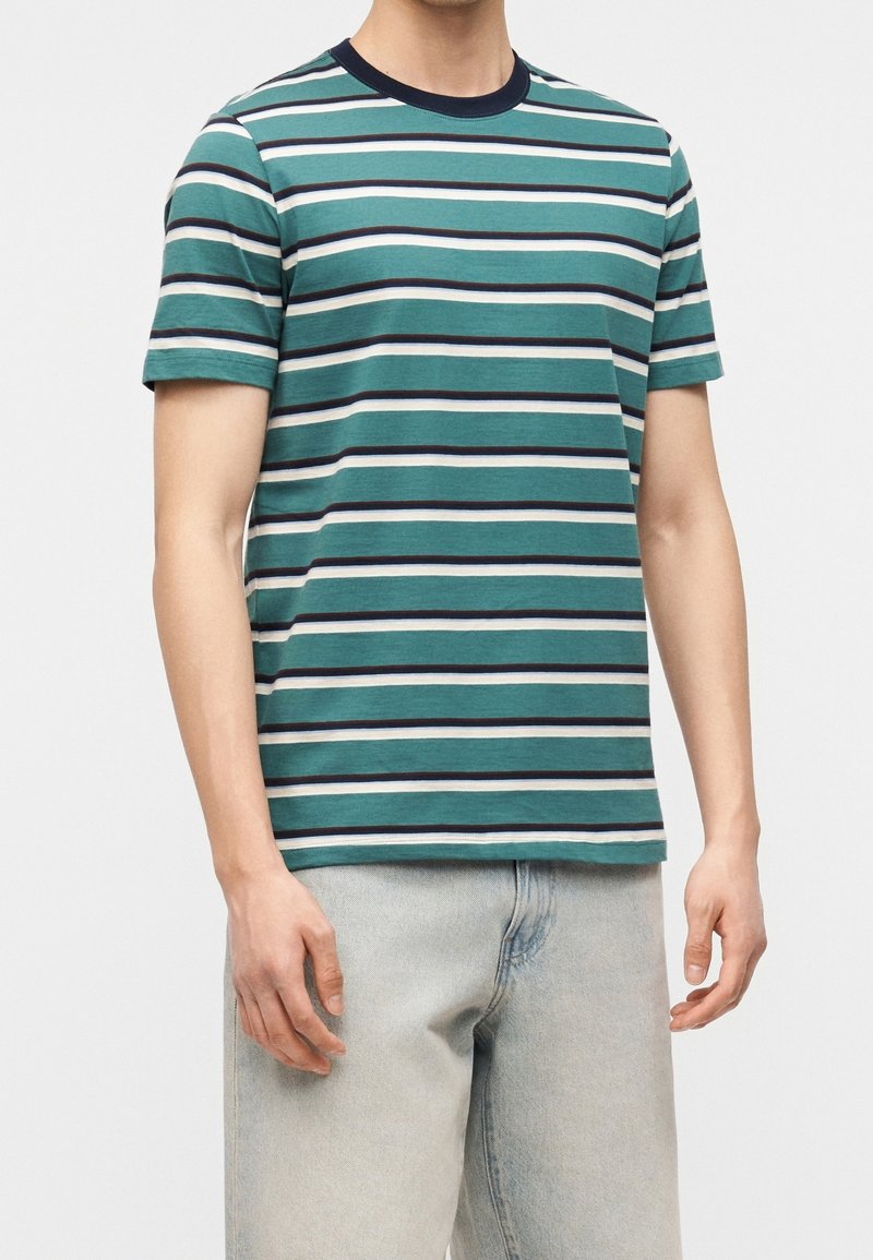 Homme portant un t-shirt à manches courtes rayé bleu sarcelle, noir et blanc avec un jean gris clair, debout devant un fond uni.