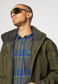 Veste en nylon verte avec doublure grise, portée par-dessus une chemise en flanelle à carreaux bleus et verts. Grande paire de lunettes de soleil rectangulaires avec verres ambrés.