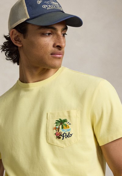 Νεαρός άνδρας με σγουρά μαλλιά, που φοράει μπλε και μπεζ καπέλο από δίχτυ και κίτρινη μπλούζα με κεντημένη τροπική σκηνή στην τσέπη του στήθους.
