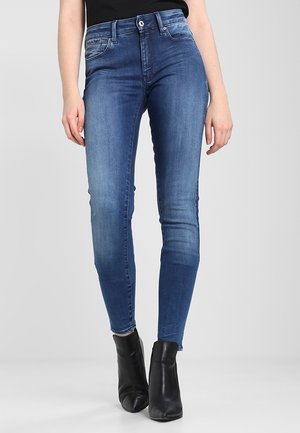 Vaqueros ajustados en denim azul oscuro; cuentan con un cierre de botón frontal, cinco bolsillos y zonas desgastadas cerca de las rodillas. Combinados con botas negras hasta el tobillo.