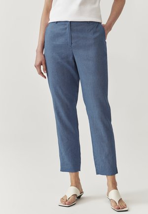 Modré kalhoty se vzorem z lehkého materiálu, s upraveným střihem, rovnými nohavicemi a čistým, minimalistickým designem. Nosené s bílými sandály.