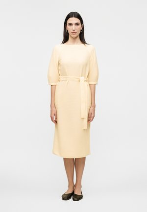 Femme aux longs cheveux noirs portant une robe midi jaune clair à manches trois-quarts avec une ceinture nouée, associée à des chaussures plates noires.