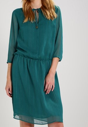 Femme portant une robe sarcelle longueur genou à rayures verticales, manches trois-quarts, avec un col noué.