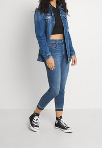 Tmavě modrá džínová bunda navrstvená přes černý crop top, spojená s modrými skinny džíny a černými vysokými teniskami s bílými podrážkami.