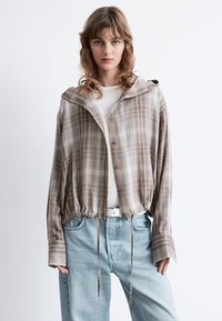 Brown plaid cropped shirt with a hood, featuring button closure, drawstring waist, and long sleeves, worn over a white top and blue jeans.
