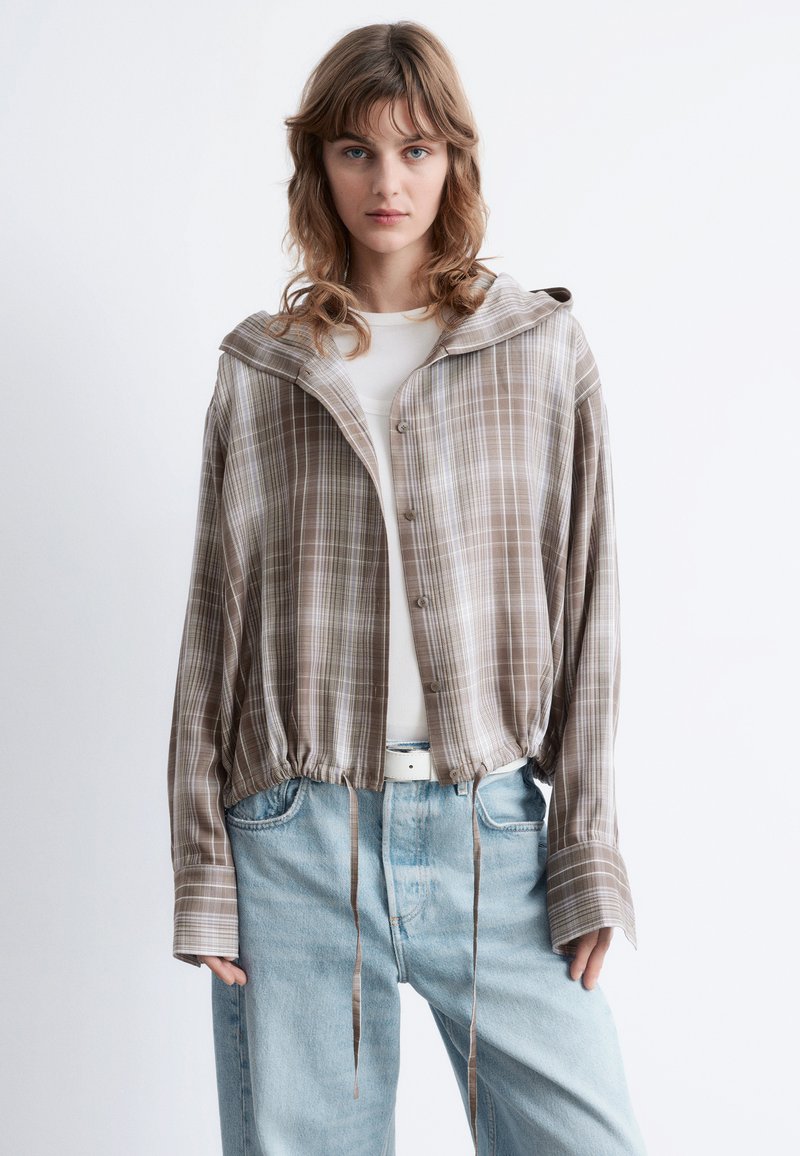 Brown plaid cropped shirt with a hood, featuring button closure, drawstring waist, and long sleeves, worn over a white top and blue jeans.