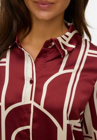 Silk skjorte med knapper i bordeaux med et cremefarvet geometrisk mønster. Har krave og en glat, skinnende overflade. To synlige knapper.