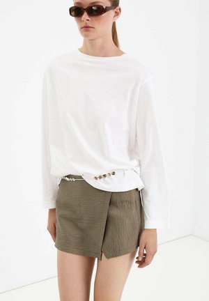 Femme portant des lunettes de soleil rectangulaires foncées, une chemise blanche à manches longues avec des boutons latéraux, et une jupe courte asymétrique marron rayée.