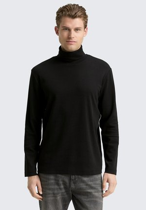 Junger Mann mit kurzen blonden Haaren, der einen schwarzen Rollkragenpullover und graue Jeans trägt, steht vor einem einfarbigen hellen Hintergrund.