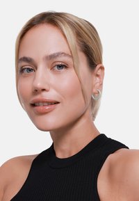 Geribbeld zwart mouwloos topje, zilveren ringoorbel, gladde huid, blauwe ogen, lichtblond haar strak naar achteren gestyled, neutrale lipglans.