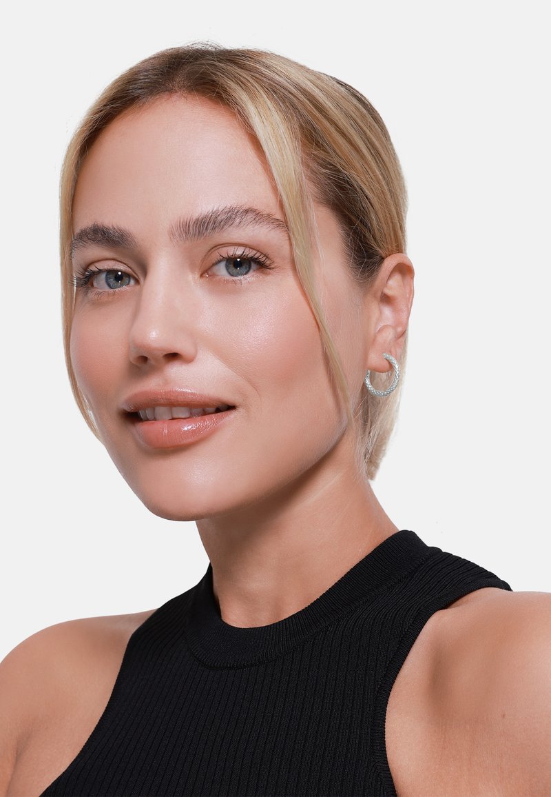 Geribbeld zwart mouwloos topje, zilveren ringoorbel, gladde huid, blauwe ogen, lichtblond haar strak naar achteren gestyled, neutrale lipglans.