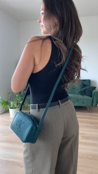 Pequena bolsa retangular cruzada de couro teal com logótipo em relevo, alça ajustável e fecho de zíper, usada com calças cinzas e uma blusa preta.