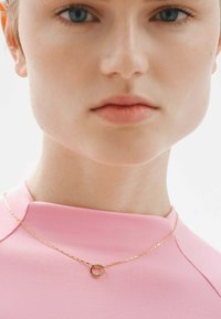 Collar de cadena de oro con un pequeño colgante circular, llevado sobre una blusa rosa de cuello alto; textura suave y diseño minimalista.