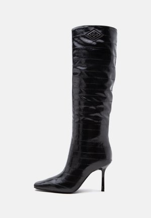 Botte noire brillante à talons hauts montant jusqu'au genou, avec un bout pointu et un petit logo en forme de diamant près du haut.