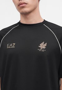 Νέος φορώντας μαύρο μπλουζάκι Emporio Armani EA7 με λογότυπο Milano Italia και άσπρες ρίγες στους ώμους σε λευκό φόντο.