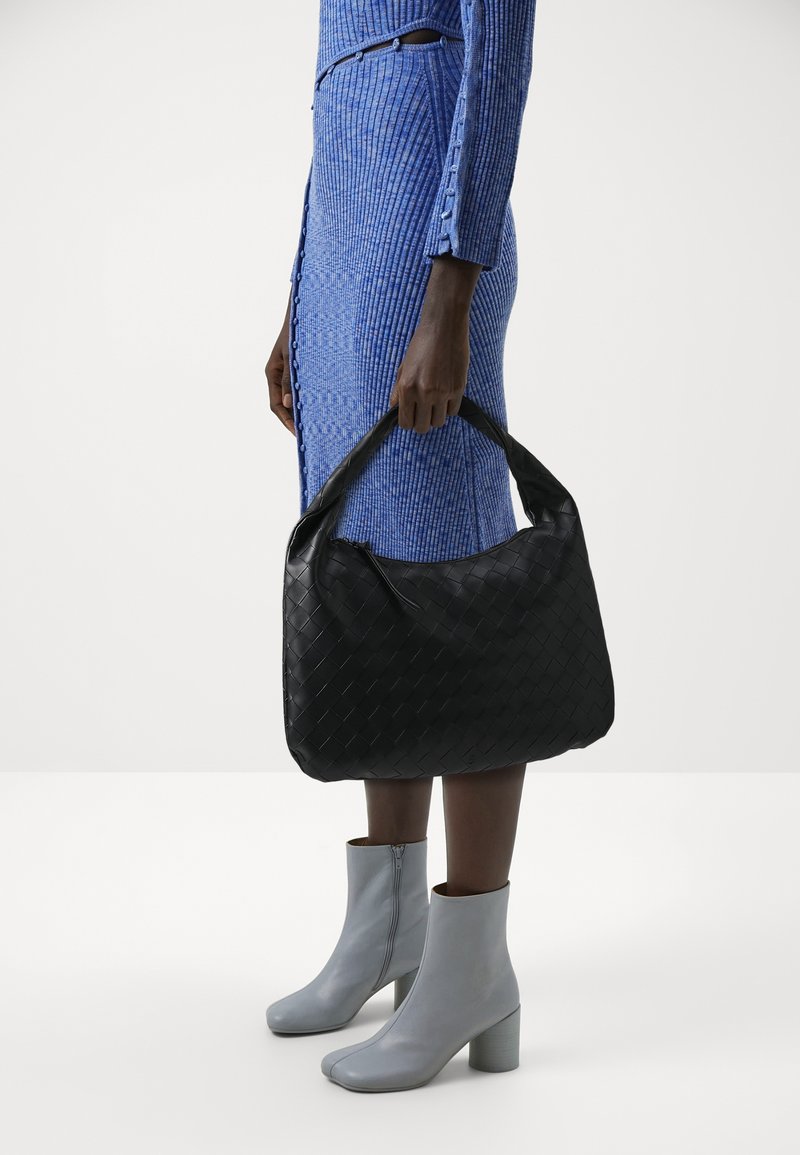 Femme en robe côtelée bleue et bottines grises claires tenant un grand sac à main noir tissé en cuir par ses anses.