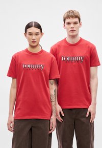Camisetas de algodón rojas con cuello redondo, con el logo "tommy" en negro y blanco, llevadas por dos modelos. Los modelos las combinan con pantalones marrones.