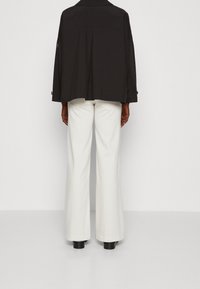 Mustat ylisuuruiset takki, jossa on laskos selässä, yhdistettyinä leveälahkeisiin valkoisiin housuihin ja mustiin saappaisiin, nähtynä takaa.