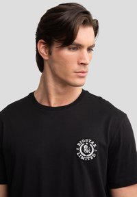 Schwarzes Baumwoll-T-Shirt mit rundem Ausschnitt und einem kleinen weißen Logo-Graphic auf der linken Brust. Glatte Textur, kurze Ärmel.