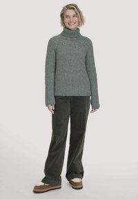 Grüner Strickrollkragenpullover mit gerippter Textur, kombiniert mit dunklen Cordhosen und braunen Schuhen mit weißen Fellbesatzakzenten.