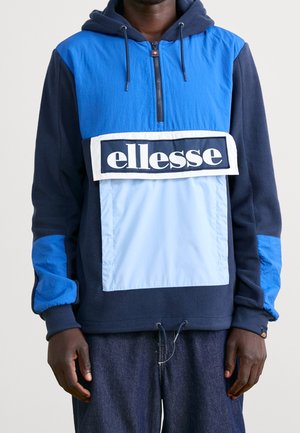 Mężczyzna w niebieskiej i granatowej bluzie z kapturem ellesse z przednią kieszenią i sznurkami, w zestawieniu z ciemnoniebieskimi spodniami, stojący na jednolitym tle.
