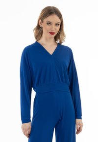 Királykék, hosszú ujjú felső V-nyakkal, illeszkedő derékrésszel és puha, sima textúrával. Elegáns szabás és egyszerű tervezési részletek.