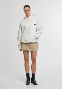 Šedá bavlněná mikina s kapucí, přední kapsou a černým logem, doplněná béžovou pletenou sukní a černými kotníkovými botami. Model stojí před jednobarevným pozadím.