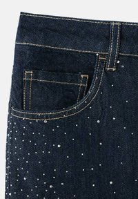 Jeans de mezclilla azul oscuro con un bolsillo frontal, decorados con tachuelas plateadas dispersas en la mitad inferior y costuras amarillas en contraste.