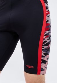 Στενά μαύρα μαγιό με κόκκινη πλευρική ρίγα και πάνελ με αφηρημένο μοτίβο, κόκκινο λογότυπο Speedo κοντά στον αριστερό μηρό, φορεμένα από άτομο.