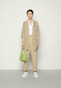 Mulher vestindo um cardigan aberto de padrão bege, top branco, calças bege, sapatilhas brancas, segurando uma bolsa verde, em pé contra uma parede lisa.