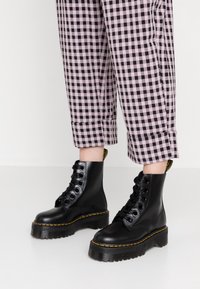 Botas de tobillo de cuero negro con suelas gruesas y texturizadas. Presentan puntera redonda, costuras amarillas y cierre con cordones en la parte delantera. Combinadas con pantalones de cuadros en rosa y negro.