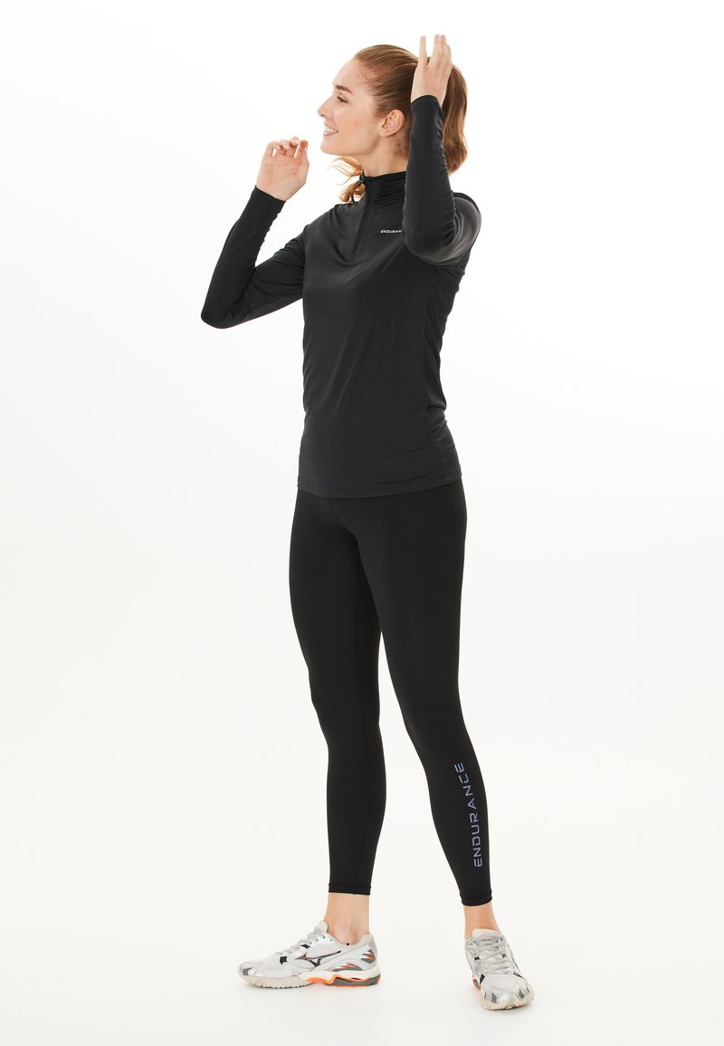 Svart långärmad sporttop med hög krage och delvis dragkedja, tillsammans med åtsittande svarta leggings med "ENDURANCE" i blått. Silverfärgade löparskor.