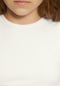 Close-up van het lagere gezicht en de nek van een persoon met lichte huid, rossig haar, gekleed in een witte geribbelde crewneck shirt.
