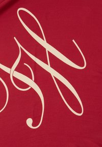 Punainen kangas esittelee suurta, kermaista, kursiivista tekstiä, jossa on laajoja viivoja ja kaaria, luoden rohkean graafisen suunnittelun.