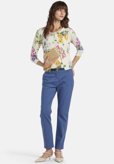 Virágmintás kardigán háromnegyedes ujjakkal, kék slim-fit nadrággal és krémszínű cipővel párosítva. A nő egy fonott borítéktáskát tart.