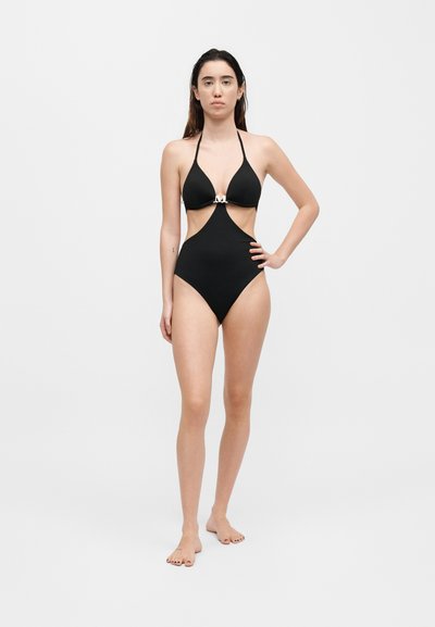 Γυναίκα στέκεται ξυπόλητη φορώντας μαύρο ολόσωμο μαγιό με ανοίγματα και λουράκια halter, με μεταλλικό κούμπωμα στο στήθος, σε λευκό φόντο.