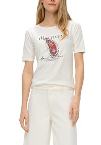 T-shirt en coton blanc à manches courtes, présentant un design graphique coloré et du texte sur le devant. Porté avec un pantalon de couleur claire.
