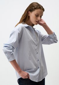 Camicia button-up azzurro chiaro con strisce verticali bianche, maniche lunghe arrotolate ai polsi, vestibilità leggermente rilassata e colletto classico.