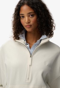 Felpa crema con zip e colletto alto, con una camicia a righe blu e bianche sotto e un piccolo logo ricamato sul petto.
