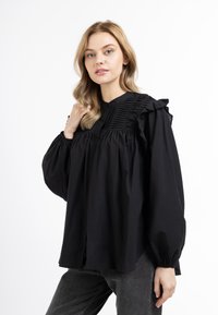 Sort langærmet bluse med puffede ærmer, volange skuldredetaljer og plisseret front. Stoffet ser blødt og let ud.